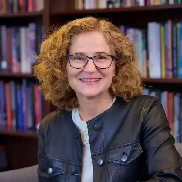 Mary Gallagher, professional woman with pale skin, red hair, and glasses wearing a white shirt and leather jacket smiles and poses in front of a wall of bookcases.